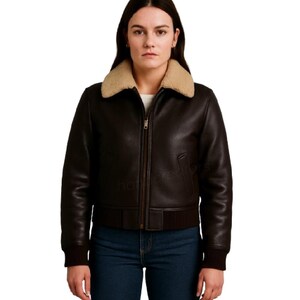 Puede incluir: Chaqueta de cuero marrón oscuro con cuello de borrego beige. La chaqueta tiene cremallera frontal, puños y dobladillo de canalé. La modelo lleva vaqueros azules. La chaqueta es de estilo bomber.