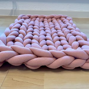 Op de afbeelding: Een vierkant, dik gebreid vloerkleed in een zachte roze tint. Het vloerkleed heeft een gevlochten rand en een dicht, getextureerd oppervlak, waardoor een gezellige en uitnodigende uitstraling ontstaat. De dikke garenconstructie suggereert een comfortabele en duurzame vloerbedekking.