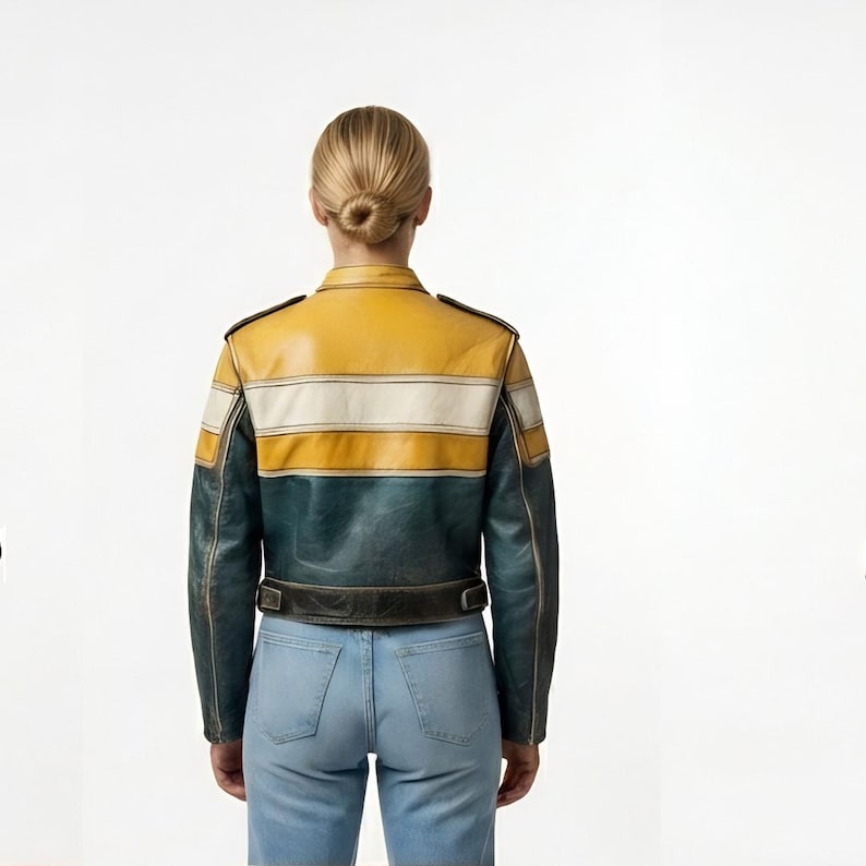 Op de afbeelding: Een leren jas met een mosterdgeel rug- en schoudergedeelte, een horizontale witte streep en een donkerblauwgroen onderste gedeelte. De jas heeft lange mouwen, een kraag en een tailleband met metalen gespen, gecombineerd met een lichtblauwe spijkerbroek.