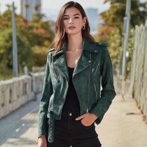 May include: A dark green suede biker jacket with a notched collar, asymmetrical zipper closure, and a belt at the waist. The jacket has long sleeves and multiple zippered pockets. The model is wearing a black top and black jeans.