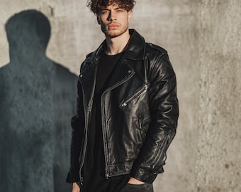 May include: A black leather motorcycle jacket with a notched collar and zippered pockets. The jacket is worn over a black t-shirt and paired with black jeans. The person is standing in front of a gray concrete wall.