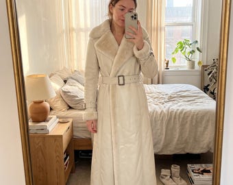 May include: A long, cream-colored coat featuring a wide faux fur collar, matching faux fur cuffs, and a belted waist with a rectangular buckle. The coat has a smooth, leather-like exterior finish and a full-length silhouette.