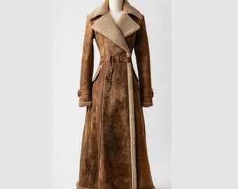 May include: A long, brown suede coat with a beige shearling collar and trim. The coat features a belted waist, long sleeves, and a flared skirt. The coat is displayed on a mannequin against a white background.