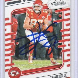 Puede incluir: Una tarjeta coleccionable de fútbol con Travis Kelce en uniforme rojo y blanco, sosteniendo un balón. La tarjeta tiene un borde rojo y blanco con las palabras "Absolute Football" en la parte superior. La firma del jugador es visible.