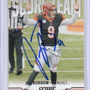 May include: A Score Team trading card featuring Joe Burrow of the Cincinnati Bengals in a black and orange uniform with the number 9, arms raised in celebration on a football field, with a blue ink autograph across the center of the card.