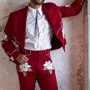 Könnte beinhalten: Ein roter Anzug im Western-Stil mit weißen Blumenstickereien und weißen Verzierungen. Der Anzug besteht aus einer Jacke und einer Hose. Die Person trägt ein weißes Hemd mit Knöpfen und eine Bolo-Krawatte. Der Anzug ist ein mutiges Mode-Statement.