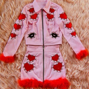 May include: A pink two-piece outfit comprising a jacket and skirt. The jacket and skirt are decorated with red heart appliqués, silver studs, and black lip details. Red feather trim adorns the cuffs and skirt hem. The outfit features a zip closure.