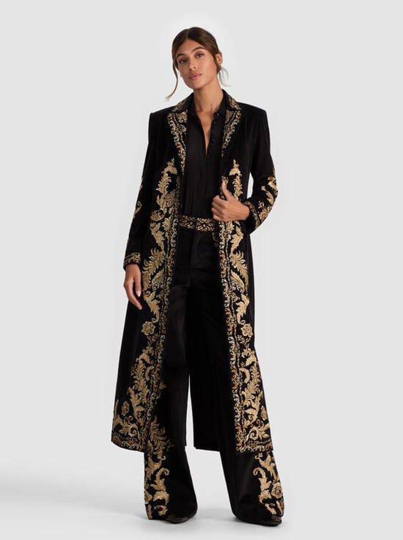 May include: A black velvet duster coat with gold embroidered floral patterns. The coat features a long, flowing design with ornate detailing along the edges and sleeves. The outfit includes matching black pants and a black shirt.
