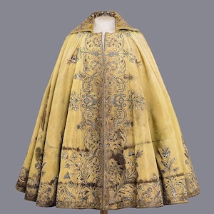 May include: A vintage yellow cape with intricate floral embroidery in shades of brown and silver. The cape features a high collar and a full, flowing design, showcasing detailed craftsmanship. The embroidery is concentrated along the front and edges.