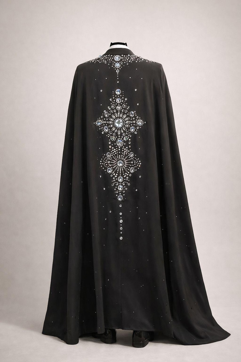 Puede incluir: Una larga capa negra adornada con brillantes pedrer&iacute;a y cristales. Los adornos forman un dise&ntilde;o sim&eacute;trico en la espalda, ofreciendo un aspecto glamuroso y elegante. La capa llega hasta el suelo.