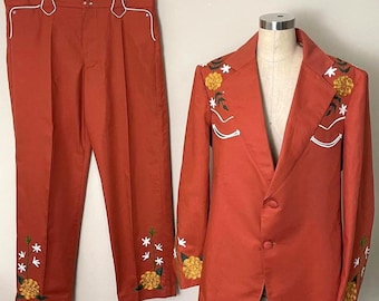 May include: Rust-colored suit set featuring a blazer and matching pants. The blazer and pants are adorned with white stitching and floral embroidery in yellow and green. The suit is displayed on a mannequin and hanger.