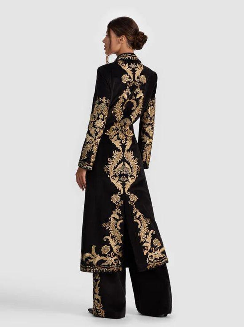May include: Black velvet coat and matching pants set with gold floral embroidery. The long coat features a high collar and a split back. The gold embroidery is on the sleeves, back, and pant legs. The outfit is elegant and suitable for special occasions.