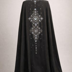 Puede incluir: Una larga capa negra adornada con brillantes pedrer&iacute;a y cristales. Los adornos forman un dise&ntilde;o sim&eacute;trico en la espalda, ofreciendo un aspecto glamuroso y elegante. La capa llega hasta el suelo.