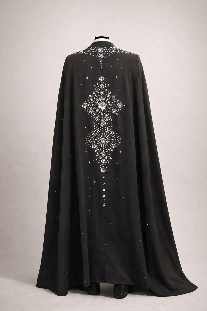 Puede incluir: Una larga capa negra adornada con brillantes pedrer&iacute;a y cristales. Los adornos forman un dise&ntilde;o sim&eacute;trico en la espalda, ofreciendo un aspecto glamuroso y elegante. La capa llega hasta el suelo.