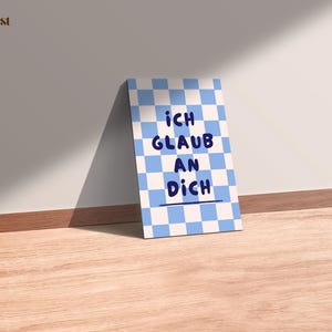 Könnte beinhalten: Eine rechteckige Karte mit blau-weißem Schachbrettmuster. Die Karte zeigt den Text "ICH GLAUB AN DICH" in Blau, vor dem Schachbrettmuster. Die Karte lehnt an einer weißen Wand auf einer Holzoberfläche.