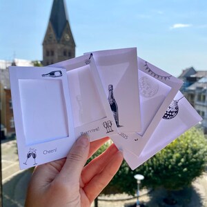 May include: A hand holds a stack of white cards with black illustrations and text. The cards feature champagne bottles, glasses, and a disco ball. Text includes "Cheers!" and "Partytime!" against a backdrop of a church steeple and blue sky.