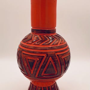 May include: A tall, orange ceramic vase with a bulbous body and a cylindrical neck. The vase features a geometric pattern in shades of brown and orange. The base is a pedestal shape. A decorative home decor item.