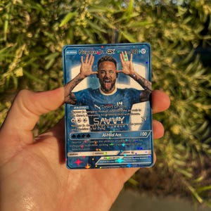 Peut inclure: Carte de collection Neymar Jr. GX bleue et argent&eacute;e avec une photo du footballeur. La carte porte les mentions "Al-Hilal Ace" et "Desert Dribble" ainsi que d'autres textes et chiffres li&eacute;s au jeu. La carte est tenue dans une main.