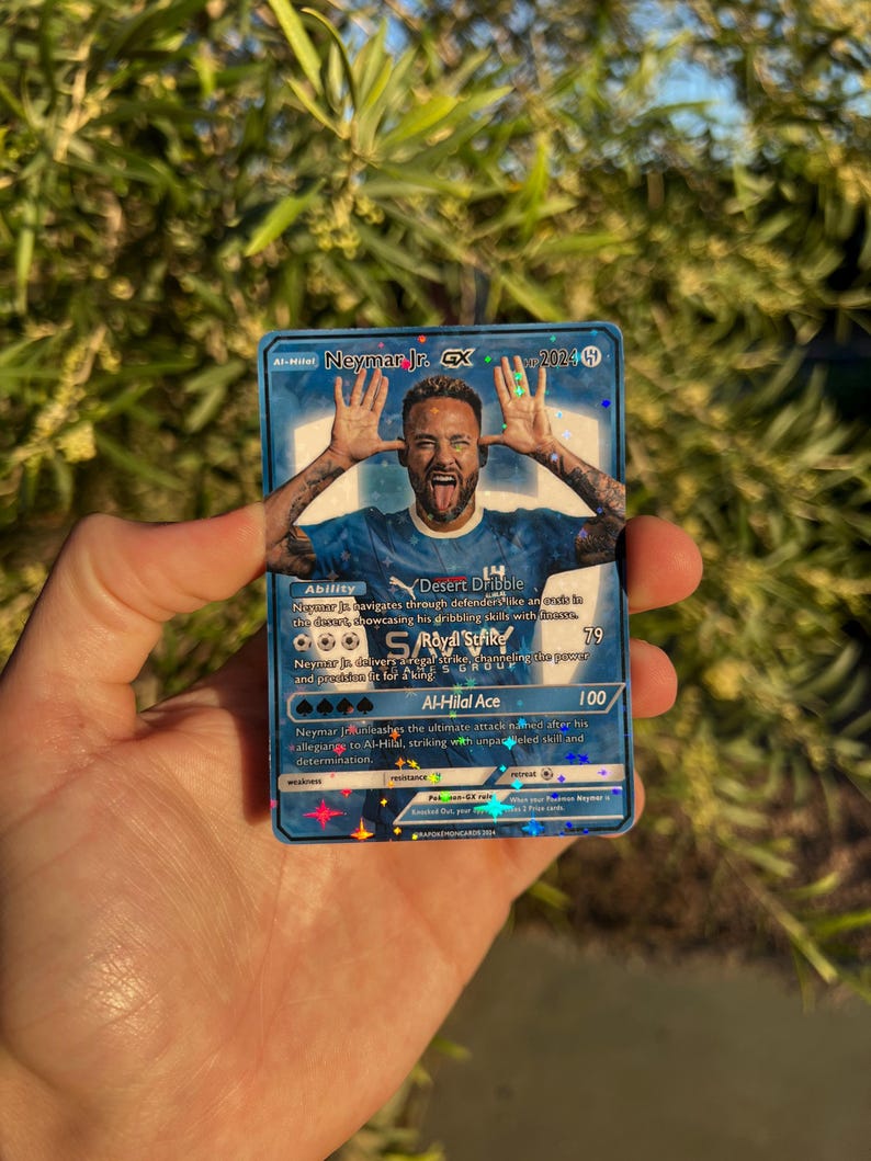 Peut inclure: Carte de collection Neymar Jr. GX bleue et argent&eacute;e avec une photo du footballeur. La carte porte les mentions "Al-Hilal Ace" et "Desert Dribble" ainsi que d'autres textes et chiffres li&eacute;s au jeu. La carte est tenue dans une main.