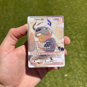 Puede incluir: Una tarjeta coleccionable holográfica Appa GX con el personaje Appa de Avatar: El último maestro del aire. La tarjeta tiene 1000 HP e incluye descripciones de ataque. La tarjeta se sostiene en una mano sobre un fondo verde.