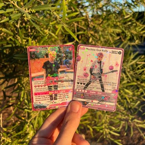 May include: Two pink Machine Gun Kelly trading cards. The cards feature images of the artist, with text including "Machine Gun Kelly 2022" and "GX". The cards are held in front of a green leafy background.