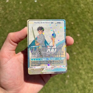 Puede incluir: Una carta de comercio de Pok&eacute;mon que presenta una garza azul y blanca con un ni&ntilde;o que sostiene una espada. La carta se llama "The Boy & the Heron GX" y tiene 999 PS. La carta tiene tres habilidades: "Portal of Hruch", "Heron's Guidance" y "Spirit of the Tower".