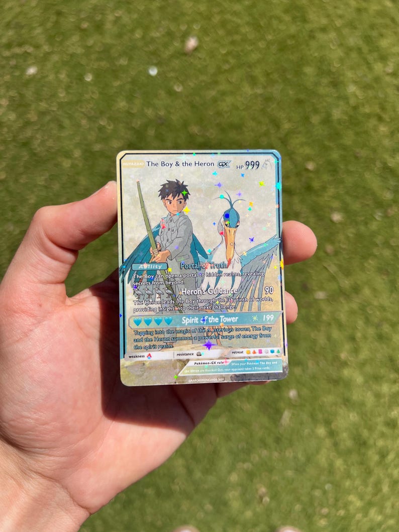 Puede incluir: Una carta de comercio de Pok&eacute;mon que presenta una garza azul y blanca con un ni&ntilde;o que sostiene una espada. La carta se llama "The Boy & the Heron GX" y tiene 999 PS. La carta tiene tres habilidades: "Portal of Hruch", "Heron's Guidance" y "Spirit of the Tower".