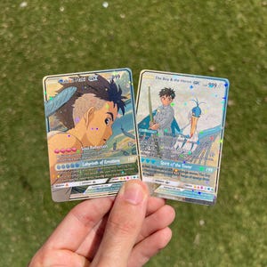 Puede incluir: Dos cartas de intercambio hologr&aacute;ficas con personajes del anime "El Ni&ntilde;o y la Garza". La carta de la izquierda presenta a Mahito Make con el texto "Reflexi&oacute;n del Alma" y "Laberinto de Emociones". La carta de la derecha presenta al Ni&ntilde;o con el texto "Gu&iacute;a de la Garza" y "Esp&iacute;ritu de la Torre".