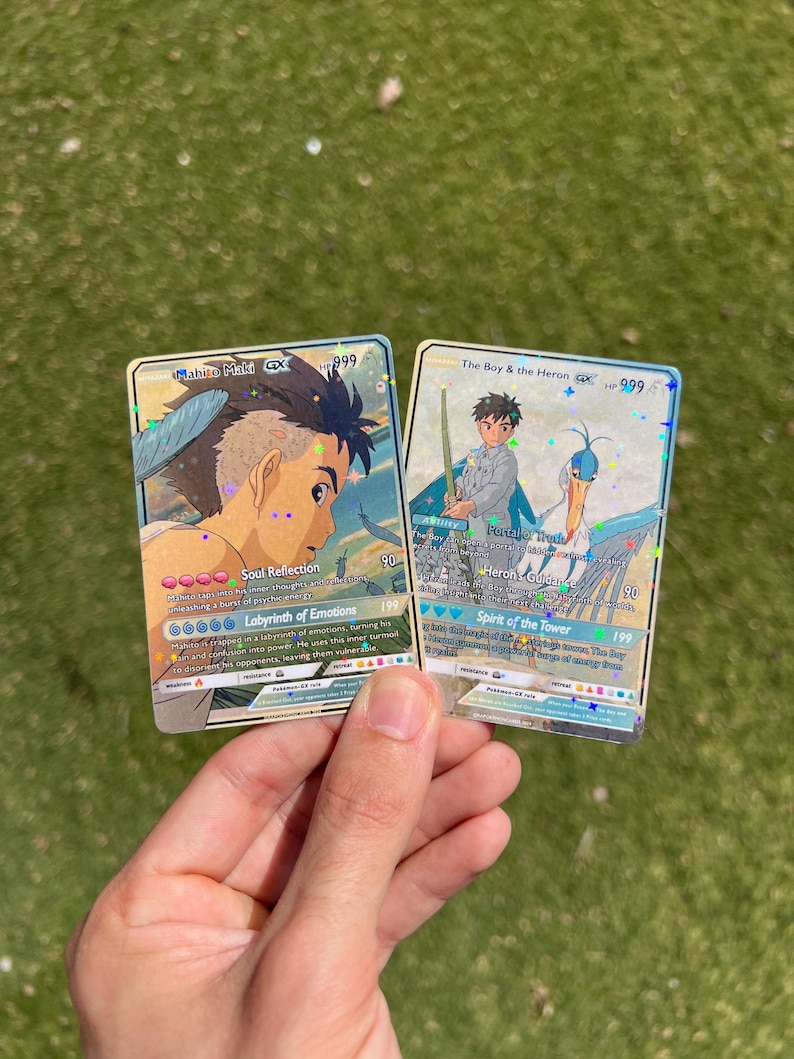 Puede incluir: Dos cartas de intercambio hologr&aacute;ficas con personajes del anime "El Ni&ntilde;o y la Garza". La carta de la izquierda presenta a Mahito Make con el texto "Reflexi&oacute;n del Alma" y "Laberinto de Emociones". La carta de la derecha presenta al Ni&ntilde;o con el texto "Gu&iacute;a de la Garza" y "Esp&iacute;ritu de la Torre".