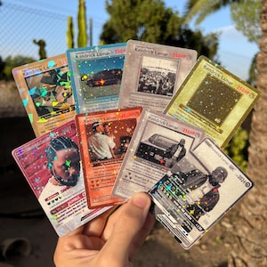 Puede incluir: Una mano sostiene una colección de cartas coleccionables con Kendrick Lamar. Las cartas tienen varios diseños, incluyendo imágenes del artista y portadas de álbumes. Las cartas tienen detalles holográficos y metálicos, con texto como "250 HP" y "HUMBLE."