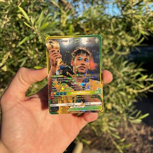 Peut inclure: Une carte &agrave; collectionner holographique repr&eacute;sentant Neymar Jr. tenant un troph&eacute;e. La carte a une bordure dor&eacute;e et verte avec le texte "Neymar Jr. GX" et "2013". La carte comprend &eacute;galement le texte "Sele&ccedil;&atilde;o Inspiration", "Samba Celebration" et "Brasil Brilliance".