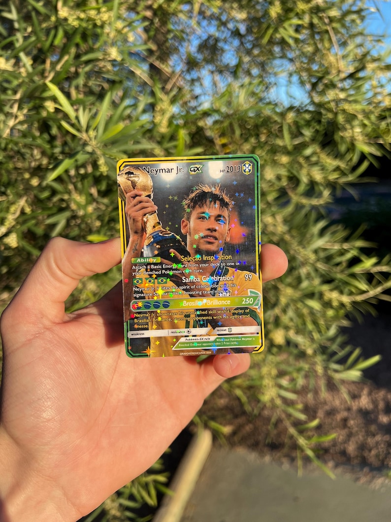 Peut inclure: Une carte &agrave; collectionner holographique repr&eacute;sentant Neymar Jr. tenant un troph&eacute;e. La carte a une bordure dor&eacute;e et verte avec le texte "Neymar Jr. GX" et "2013". La carte comprend &eacute;galement le texte "Sele&ccedil;&atilde;o Inspiration", "Samba Celebration" et "Brasil Brilliance".