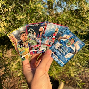 Peut inclure: Une main tient cinq cartes &agrave; collectionner Neymar Jr. Les cartes pr&eacute;sentent des images color&eacute;es de Neymar Jr. en action, avec du texte tel que "Neymar Jr. GX" et "All Ace". L'arri&egrave;re-plan est un feuillage vert flou.