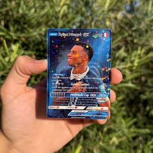 Può includere: Una carta collezionabile rettangolare con uno sfondo blu olografico e motivi a stella. La carta mostra un ritratto di Kylian Mbappé, il testo Kylian Mbappé GX, FIFA World Cup GX 2026 e varie statistiche di gioco tra cui Ability Uncatchable.