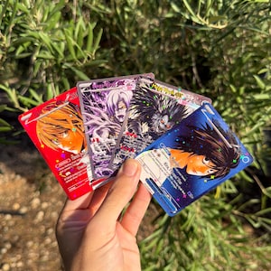 May include: A set of four rectangular trading cards featuring anime-style character illustrations. The cards display vibrant red, purple, black, and blue backgrounds with holographic star patterns, text, and character names including Light Yagami, Ryuk, and Ryuzaki.