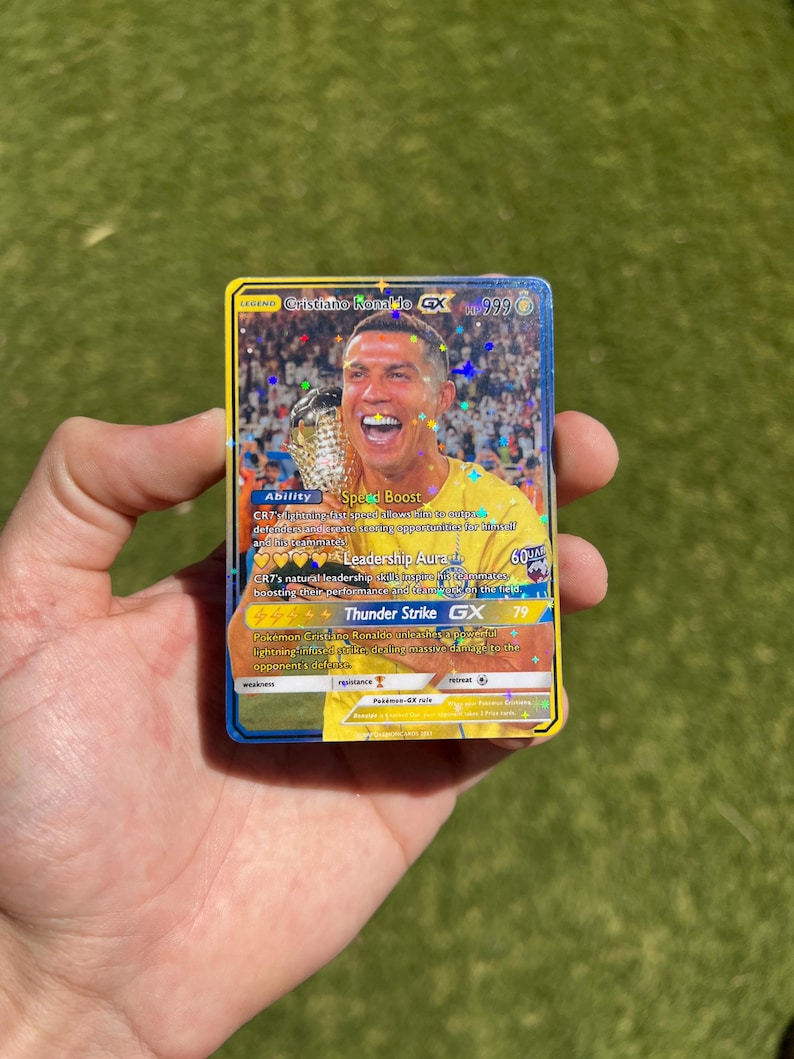May include: A Pokemon trading card featuring Cristiano Ronaldo, a soccer player, with a gold and blue border. The card is a holographic card with a shiny finish. The card's name is "Cristiano Ronaldo GX" and the card number is "999". The card has an ability called "Speed Boost" and a move called "Thunder Strike GX".