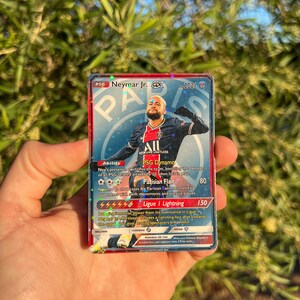 Peut inclure: Une carte &agrave; collectionner Neymar Jr. GX avec le logo PSG et l'image du joueur. La carte contient du texte d&eacute;taillant les capacit&eacute;s et les statistiques, avec un copyright 2021. La carte est tenue dans une main.