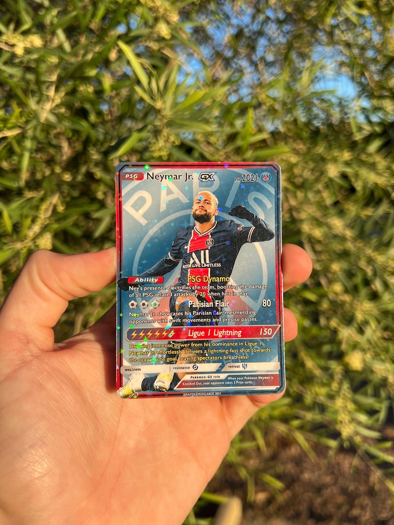 Peut inclure: Une carte &agrave; collectionner Neymar Jr. GX avec le logo PSG et l'image du joueur. La carte contient du texte d&eacute;taillant les capacit&eacute;s et les statistiques, avec un copyright 2021. La carte est tenue dans une main.