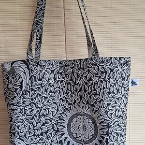 Op de afbeelding: Zwarte en witte draagtas met een bloemen- en geometrisch patroon. De tas heeft lange hengsels en een rechthoekige vorm. Het ontwerp bevat een centraal zonmotief en ingewikkelde blad- en bloemdetails. De tas is gemaakt van een stevige stof.