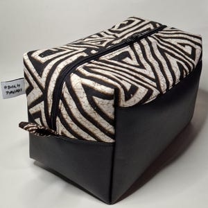 Puede incluir: Un neceser rectangular con cremallera negra. La parte superior presenta un diseño geométrico en blanco y negro, mientras que la parte inferior es negra lisa. La etiqueta dice "Batik by PAVANI".