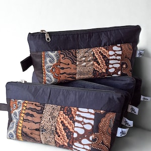 Pode incluir: Três bolsas pretas com zíper e design de patchwork. O patchwork apresenta vários padrões batik marrons e creme. Cada bolsa tem uma pequena alça preta e um zíper prateado. Etiquetas brancas são anexadas na lateral.