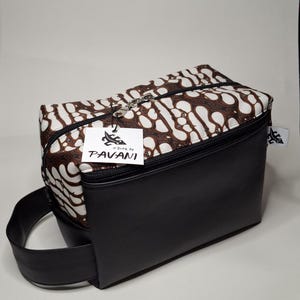 Op de afbeelding: Een rechthoekige zwarte en bruine toilettas met ritssluiting. De bovenkant van de tas heeft een bruin en wit batikpatroon, terwijl de onderkant effen zwart is. De tas heeft een zwart handvat en een label met "Pavani".