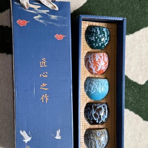 May include: A set of five small, colourful ceramic cups in a blue gift box. The cups are in shades of green, orange, blue, dark brown, and tan. The blue box has Chinese characters and illustrations of clouds and birds.