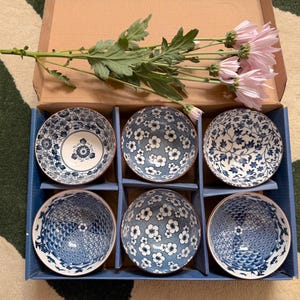 Può includere: Un set di sei ciotole in ceramica blu e bianca con motivi floreali, disposte in una scatola di cartone blu. Un rametto di fiori rosa e foglie verdi è posto sopra.
