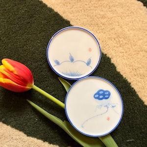 Puede incluir: Dos platos de cerámica blanca con bordes azules y delicados diseños florales. Un plato presenta una flor de loto azul, el otro tiene hojas azules. Un tulipán rojo y amarillo y hojas verdes en primer plano.