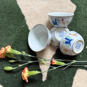 May include: Four white ceramic bowls with blue and red rooster designs. The bowls are arranged on a green and beige patterned surface, with a sprig of orange and yellow carnations in the foreground.