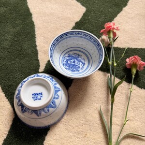 May include: Two white and blue porcelain bowls with a traditional Chinese design. The bowls feature a blue dragon motif and decorative patterns. The bowls are on a patterned rug with pink flowers.