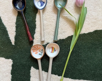May include: Five ceramic spoons with different designs. The spoons are white with blue, green, orange, and rainbow designs. The spoons are arranged in a semi-circle on a white rug with a green background.