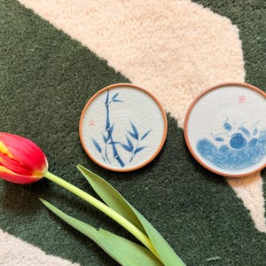 May include: Two circular ceramic coasters with blue and white bamboo and water designs, featuring reddish-brown rims. A red and yellow tulip with green leaves is in the foreground, set against a green and beige patterned background.
