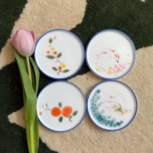 May include: Four white ceramic coasters with blue trim and hand-painted designs. The designs include a branch with orange and yellow fruit, a pink and red fish, a green and blue dragon, and a branch with orange fruit.